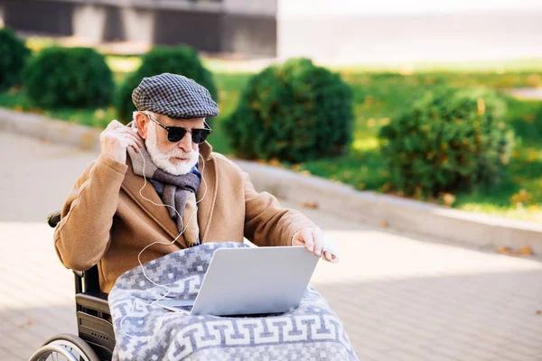 Senior behinderter Mann im Rollstuhl mit Laptop und Kopfhörer auf der Straße — Stockfoto