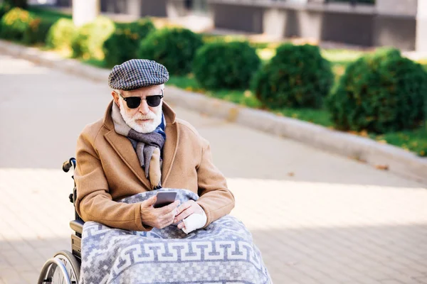 Hombre discapacitado senior en silla de ruedas usando teléfono inteligente en la calle - foto de stock