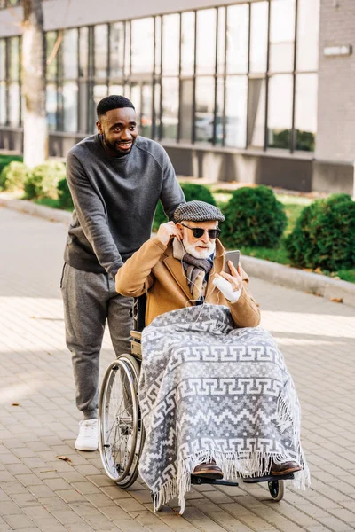 Senior disabled man in wheelchair listening musing with smartphone and earphones with african american cuidador on street — Stock Photo