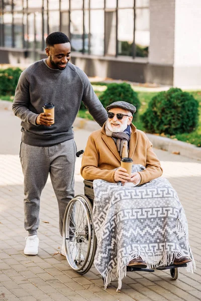 Smiling senior disabled man in wheelchair with plaid and african american man riding by street with paper cups of coffee — Stock Photo