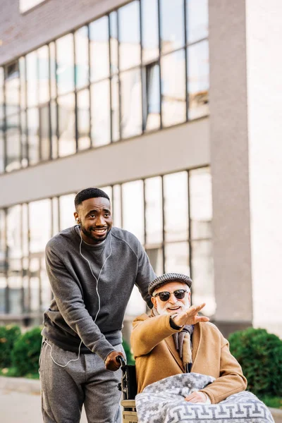 Happy senior disabled man in wheelchair with plaid and african american man riding by street and looking somewhere — Stock Photo