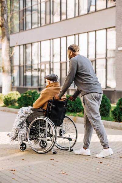 Senior disabled man in wheelchair with plaid and cuidador riding by street — Stock Photo