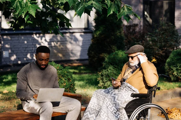 Senior discapacitado hombre en silla de ruedas y joven afroamericano hombre usando gadgets juntos en la calle — Stock Photo