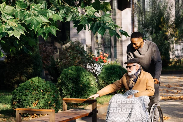 Happy senior disabled man in wheelchair and african american cuidador spending time together on street — Stock Photo