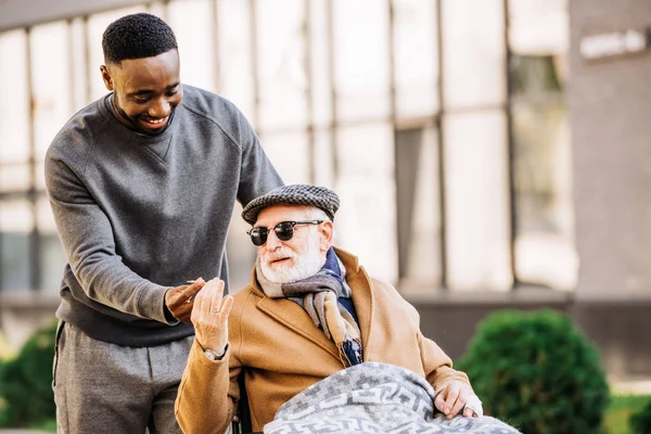 Africano americano hombre dando conjunto a senior discapacitado hombre en silla de ruedas wjhile ellos pasar tiempo juntos en calle - foto de stock