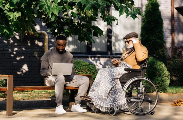 Senior disabled man in wheelchair and african american man using gadgets together on street — Stock Photo