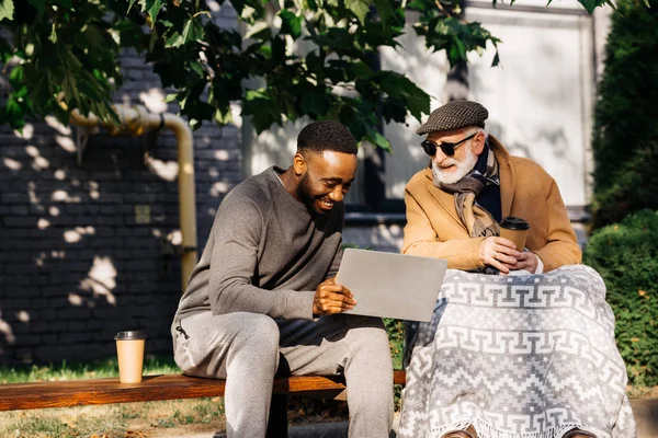 Senior disabled man in wheelchair and african american man using laptop together on street — Stock Photo