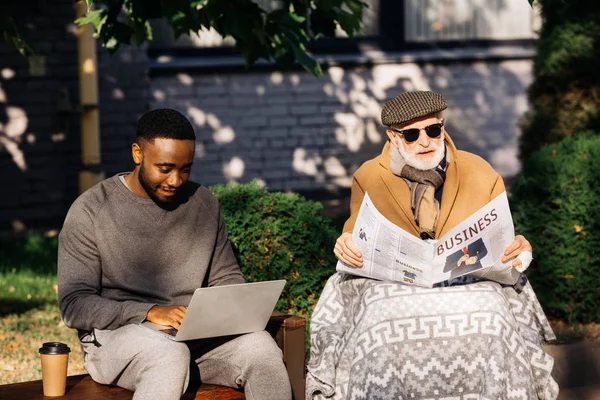 Senior disabled man reading business newspaper in wheelchair while african american man using laptop on street — Stock Photo