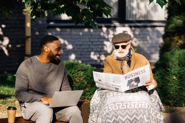 Homem deficiente sênior feliz lendo jornal de negócios em cadeira de rodas enquanto homem americano africano usando laptop na rua — Fotografia de Stock