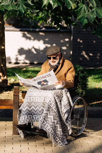 Anciano discapacitado hombre en silla de ruedas leyendo periódico de negocios en la calle - foto de stock