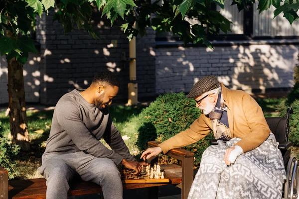 Anciano discapacitado en silla de ruedas y enfermera afroamericana jugando ajedrez juntos en la calle — Stock Photo
