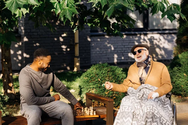 Senior disabled man in wheelchair and african american cuidador playing chess together on street — Stock Photo