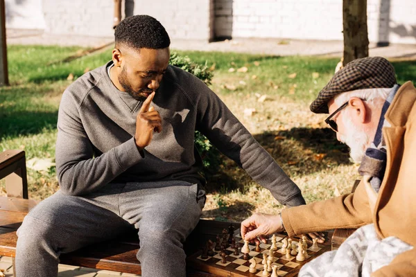 Hombre mayor y hombre afroamericano reflexivo jugando ajedrez juntos en la calle - foto de stock