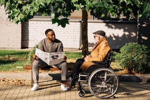 Senior handicapé en fauteuil roulant et homme afro-américain passer du temps ensemble et lire le journal dans la rue — Photo de stock