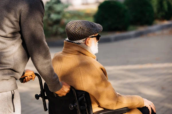 Обрезанный снимок пожилого инвалида в инвалидной коляске и медсестры, едущей по улице — стоковое фото