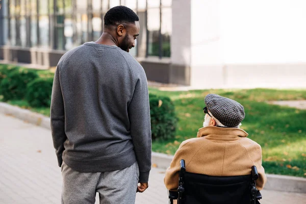 Vista trasera del anciano discapacitado en silla de ruedas y sonriente hombre afroamericano pasando tiempo juntos en la calle y mirándose el uno al otro - foto de stock