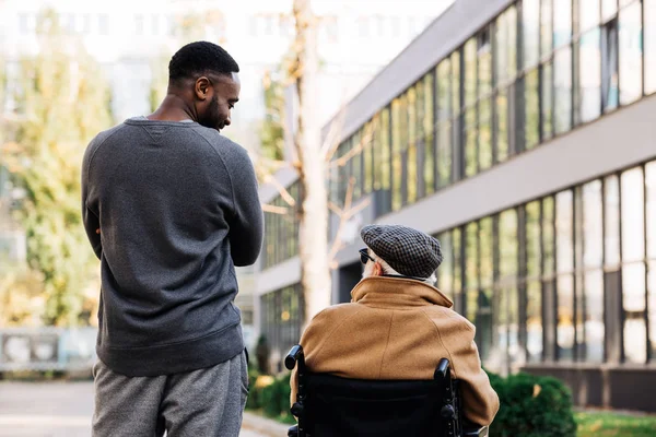 Vista trasera del hombre discapacitado sénior en silla de ruedas y hombre afroamericano pasando tiempo juntos en la calle y mirándose el uno al otro - foto de stock
