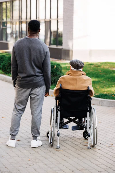 Rear view of senior disabled man in wheelchair and young african american cuidador spending time together on street — Stock Photo