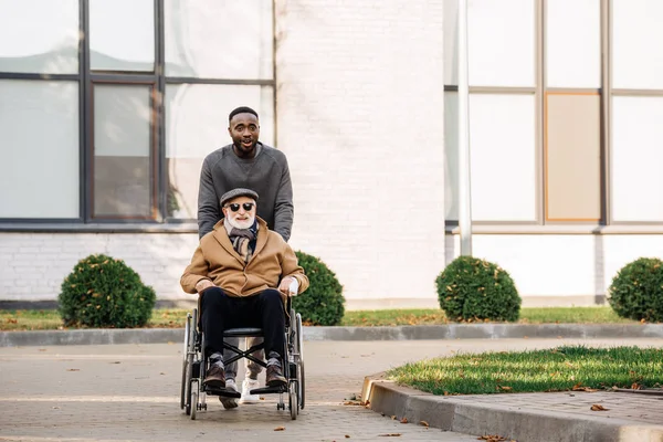 Happy senior disabled man in wheelchair and african american man riding by street together — Stock Photo