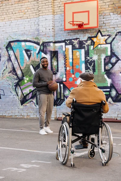 Vue arrière de l'homme handicapé âgé en fauteuil roulant regardant l'homme afro-américain jouer au basket-ball dans la rue — Photo de stock