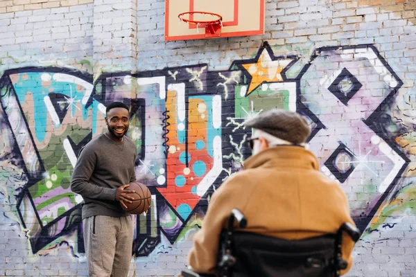 Visão traseira do homem com deficiência sênior em cadeira de rodas olhando para o homem americano africano jogando basquete na rua — Fotografia de Stock