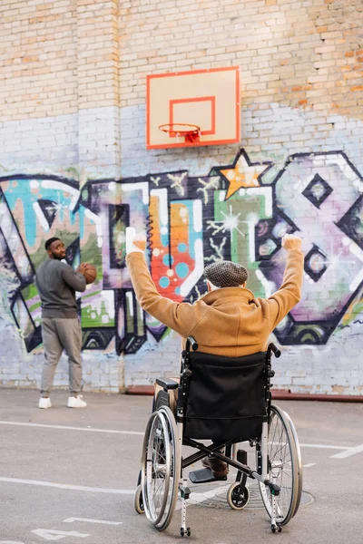 Back view of celebrating senior disabled man in wheelchair looking at african american man playing basketball on street — Stock Photo