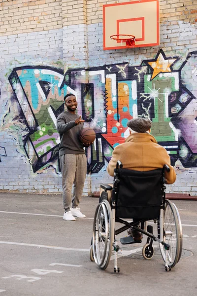 Задний вид пожилого инвалида в инвалидной коляске, смотрящего на счастливого африканского американца, играющего в баскетбол на улице — стоковое фото