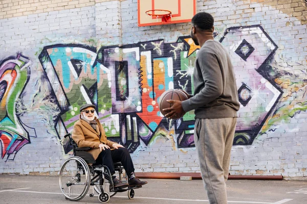 Heureux homme handicapé senior en fauteuil roulant et homme afro-américain jouer au basket-ball ensemble dans la rue — Photo de stock