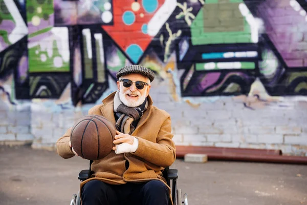 Старший инвалид в инвалидной коляске с баскетбольным мячом, смотрящий в камеру на улице — стоковое фото