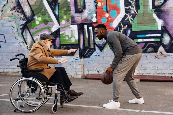 Senior discapacitado hombre en silla de ruedas y afroamericano hombre jugando baloncesto juntos en la calle - foto de stock