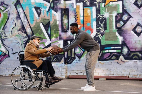 Side view of senior disabled man in wheelchair and african american man playing basketball together on street — Stock Photo