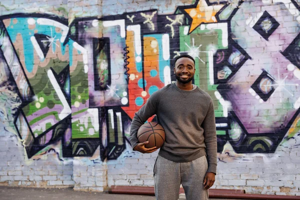 Красивый африканский американец с баскетбольным мячом смотрит в камеру на улице перед красочными граффити — стоковое фото