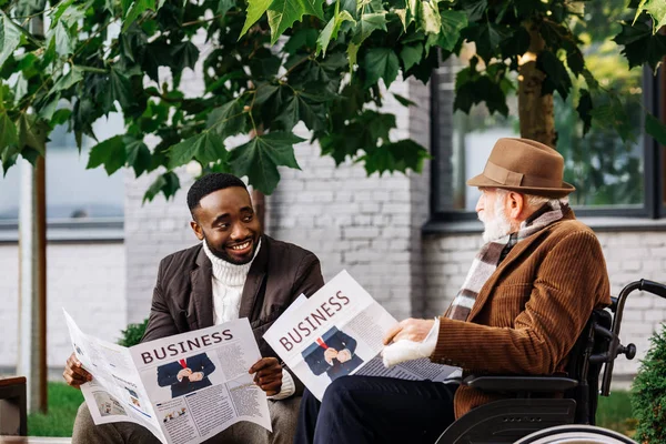Homem deficiente sênior feliz na cadeira de rodas e homem americano africano lendo jornais juntos na rua — Fotografia de Stock