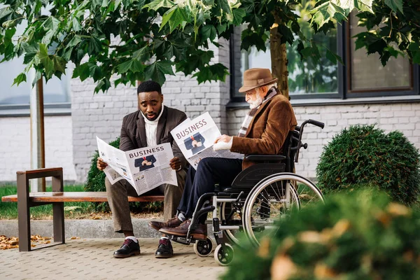 Senior disabled man in wheelchair and african american man reading newspapers together on street — Stock Photo