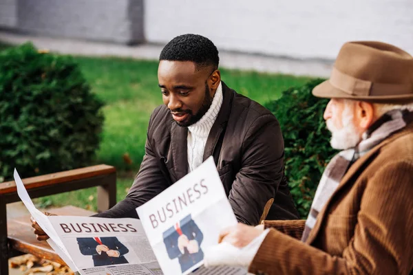 Hombre mayor y hombre afroamericano leyendo periódicos juntos en la calle - foto de stock