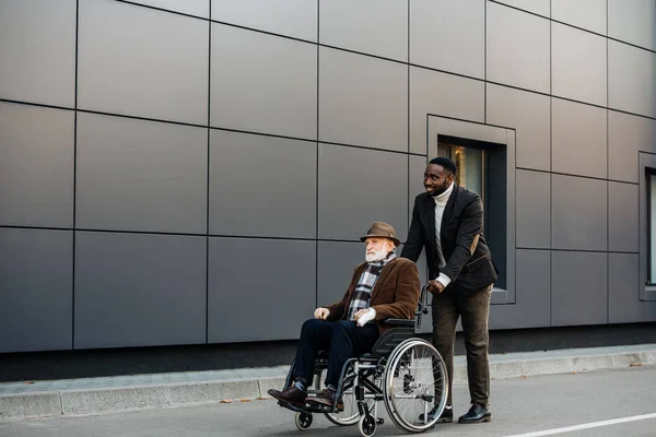 Senior disabled man in wheelchair and young african american cuidador riding by street — Stock Photo