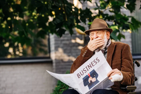 Bostezando hombre discapacitado senior en silla de ruedas leyendo periódico de negocios en la calle - foto de stock