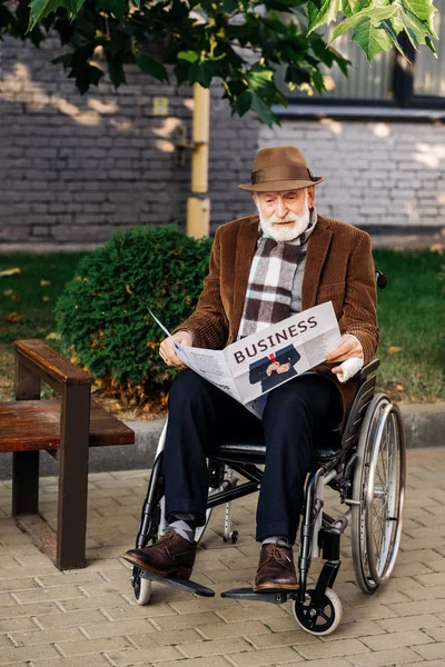 Apuesto hombre discapacitado senior en silla de ruedas leer periódico de negocios en la calle - foto de stock