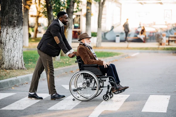 Вид сбоку пожилого инвалида в инвалидной коляске и молодых африканцев, катающихся на велосипеде по улице — стоковое фото