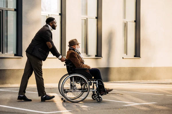 Side view of senior disabled man in wheelchair and handsome african american cuidador riding by street — Stock Photo
