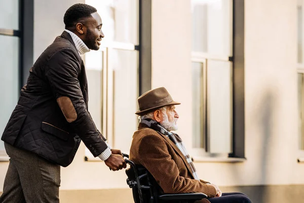 Vue latérale de l'homme handicapé âgé en fauteuil roulant et cuidador afro-américain chevauchant par la rue — Photo de stock