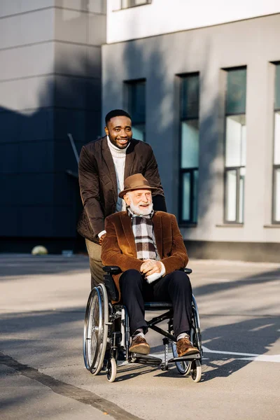 Smiling senior disabled man in wheelchair and african american cuidador riding by street — Stock Photo