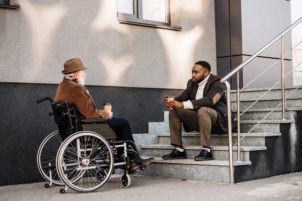 Anciano discapacitado en silla de ruedas y cuidador afroamericano tomando café juntos y charlando en la calle - foto de stock