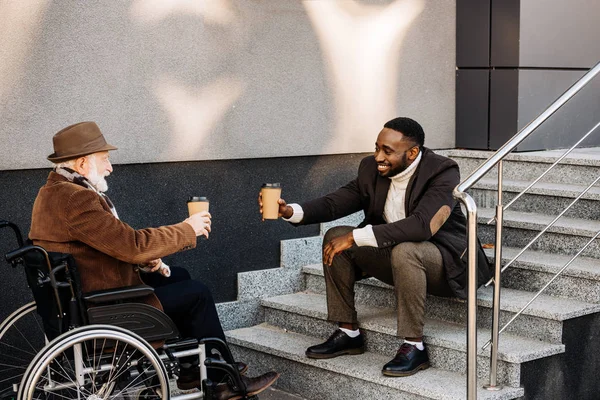 Feliz anciano discapacitado hombre en silla de ruedas y hombre afroamericano beber café juntos y tazas de papel tintineo en la calle - foto de stock