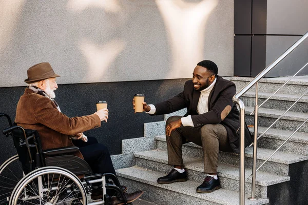 Feliz anciano discapacitado hombre en silla de ruedas y hombre afroamericano beber café juntos y tazas de papel tintineo en la calle — Stock Photo