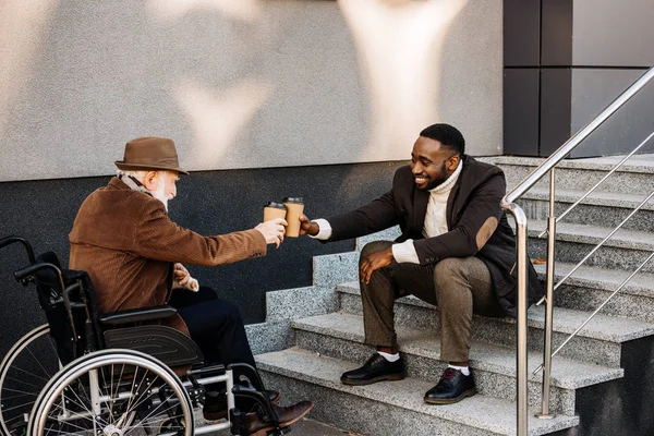 Senior disabled man in wheelchair and african american man drinking coffee together and clinking paper cups on street — Stock Photo