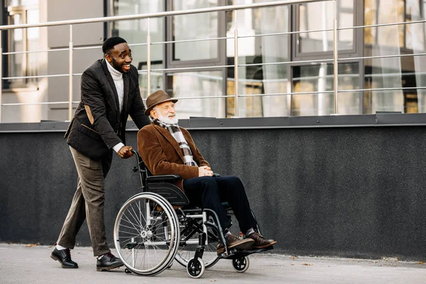 Happy senior disabled man in wheelchair and african american cuidador riding by street — Stock Photo