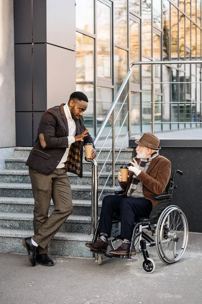 Senior disabled man in wheelchair and african american man drinking coffee together on street — Stock Photo