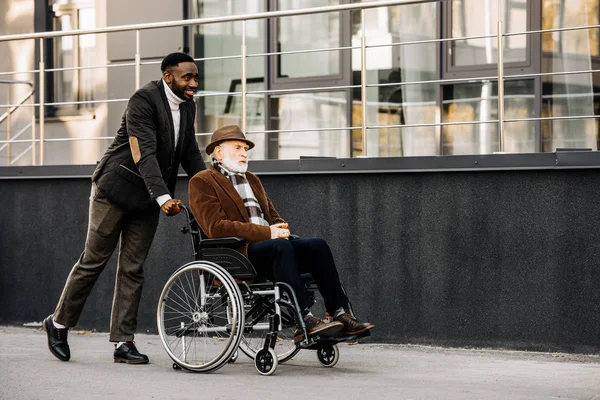 Smiling senior disabled man in wheelchair and african american man riding by street — Stock Photo