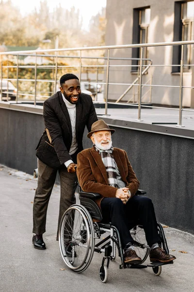 Happy senior disabled man in wheelchair and african american man riding by street — Stock Photo